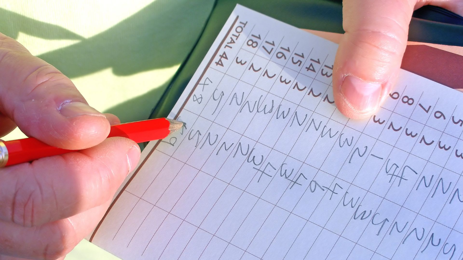 A person is marking a scorecard with a red pencil, carefully noting the golf scoring terms amid rows and columns of numbers and letters.